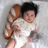 A baby with thick dark hair, in a cream onesie, rests on a blanket holding a blue toy and pacifier, lying against the plush Tummy Time Baguette pillow—ideal for tummy time and imaginative play.