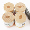 The Yogurt Play Food Pack includes four containers with wooden lids, each featuring emblems of raspberry, vanilla, strawberry, and peach. Ideal for plastic-free role play. "Yogurt" is printed on them. A black stone with a leaf design is also included.