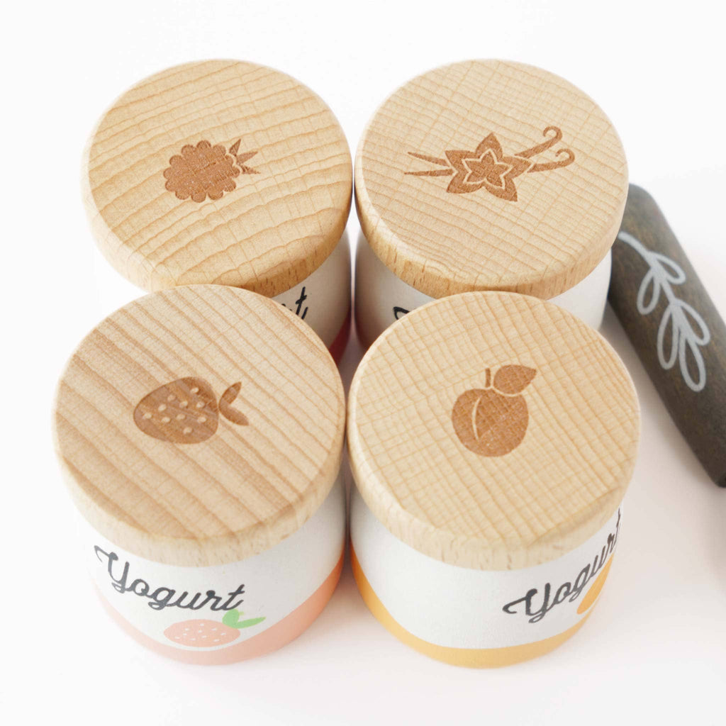 The Yogurt Play Food Pack includes four containers with wooden lids, each featuring emblems of raspberry, vanilla, strawberry, and peach. Ideal for plastic-free role play. "Yogurt" is printed on them. A black stone with a leaf design is also included.