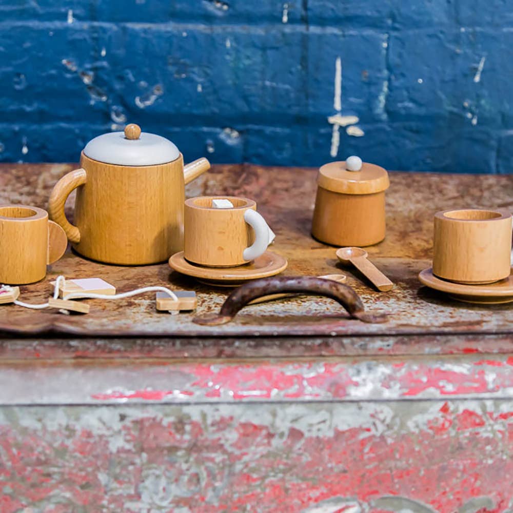A Wooden Tea Set with a teapot, two saucers with cups, a lidded sugar bowl, and a spoon is displayed on a weathered metal surface, paired with designer coffee table books against a textured blue brick wall.
