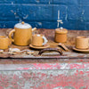 A Wooden Tea Set with a teapot, two saucers with cups, a lidded sugar bowl, and a spoon is displayed on a weathered metal surface, paired with designer coffee table books against a textured blue brick wall.