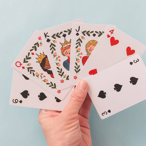 A hand holds six cards from the Sky Blue Library Card Deck Of Playing Cards—queen, king, and jack of hearts (with illustrated faces), ace of hearts, three and six of spades—against a light blue background; ships in about one week.