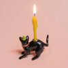 The Black Cat Cake Topper, featuring lifelike details and a green and yellow striped party hat, supports a lit yellow candle on its back. It's the ideal addition for cat-themed parties, set against a soft pink background.