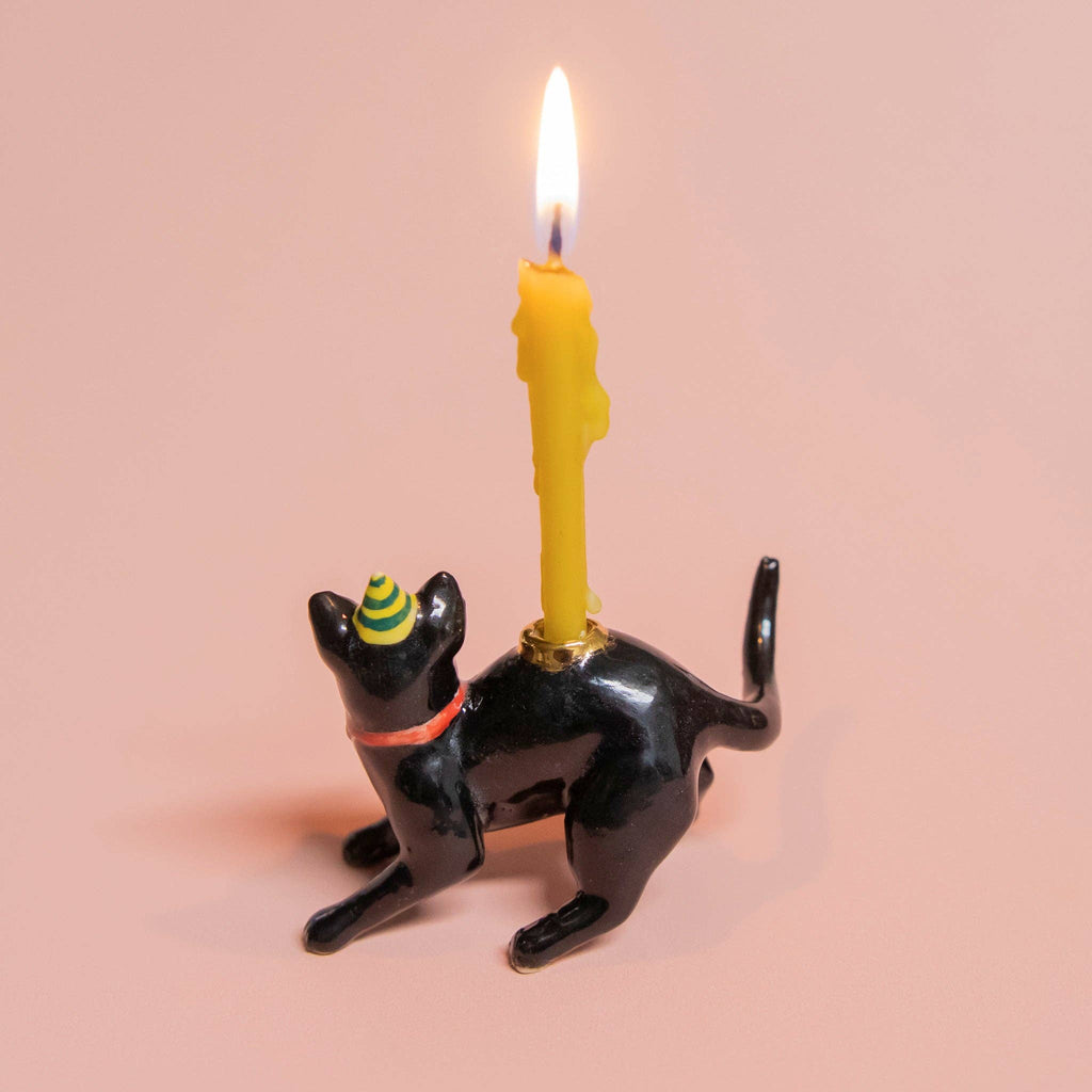 The Black Cat Cake Topper, featuring lifelike details and a green and yellow striped party hat, supports a lit yellow candle on its back. It's the ideal addition for cat-themed parties, set against a soft pink background.