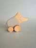 The Pull and Push Toy Little Wild Boar, handcrafted from sustainable materials, rests on a gray surface. It features a stylized animal with a curved body adorned with light brown stripes and has a smiling face, two eyes, and pink cheeks painted on it.
