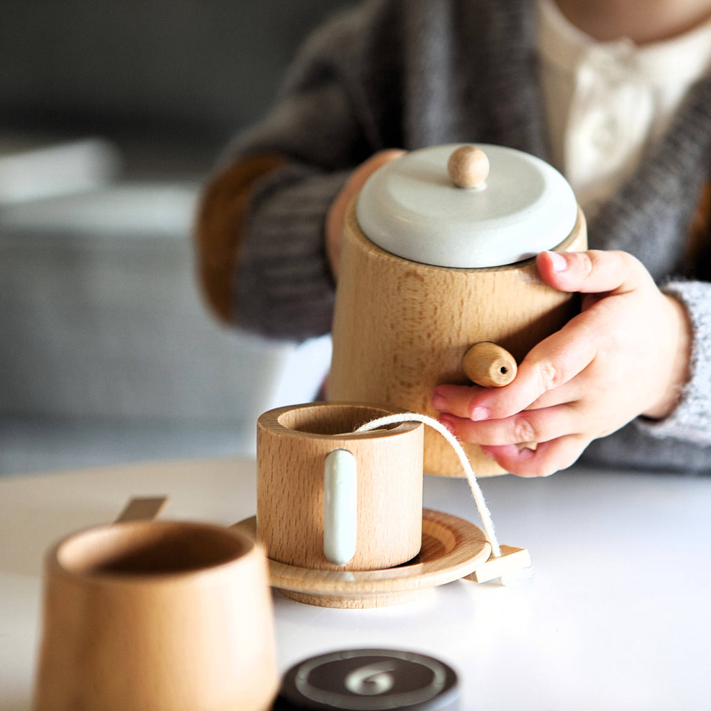 A child joyfully plays with a Wooden Tea Set, gracefully holding a teapot and teacup with a teabag inside. Sporting a gray sweater against a softly blurred background, the scene evokes the cozy warmth of an almond chai.