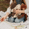 A child in a white sweater and blue jeans sits on a blanket outdoors, cuddling three Harey the Bunny plush toys with embroidered features. An open book lies nearby, suggesting a cozy reading time with their vintage-look companions.