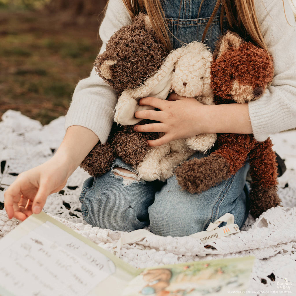 A child in a white sweater and blue jeans sits on a blanket outdoors, cuddling three Harey the Bunny plush toys with embroidered features. An open book lies nearby, suggesting a cozy reading time with their vintage-look companions.