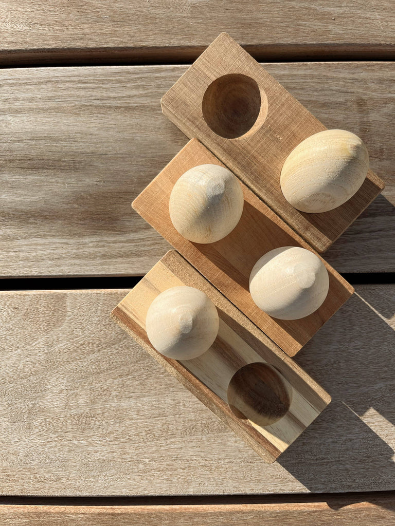 Three Wooden Egg Shakers with Stand are displayed diagonally on a wooden surface in bright sunlight. Ideal for promoting developmental milestones through tactile play.