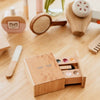 The Wooden Beauty Kit, a small play set on a wooden surface, features lipstick, an eyeshadow palette, and other cosmetic wonders. A decorative box with a sleeping face holds the timeless toy makeup, all painted with non-toxic paint.