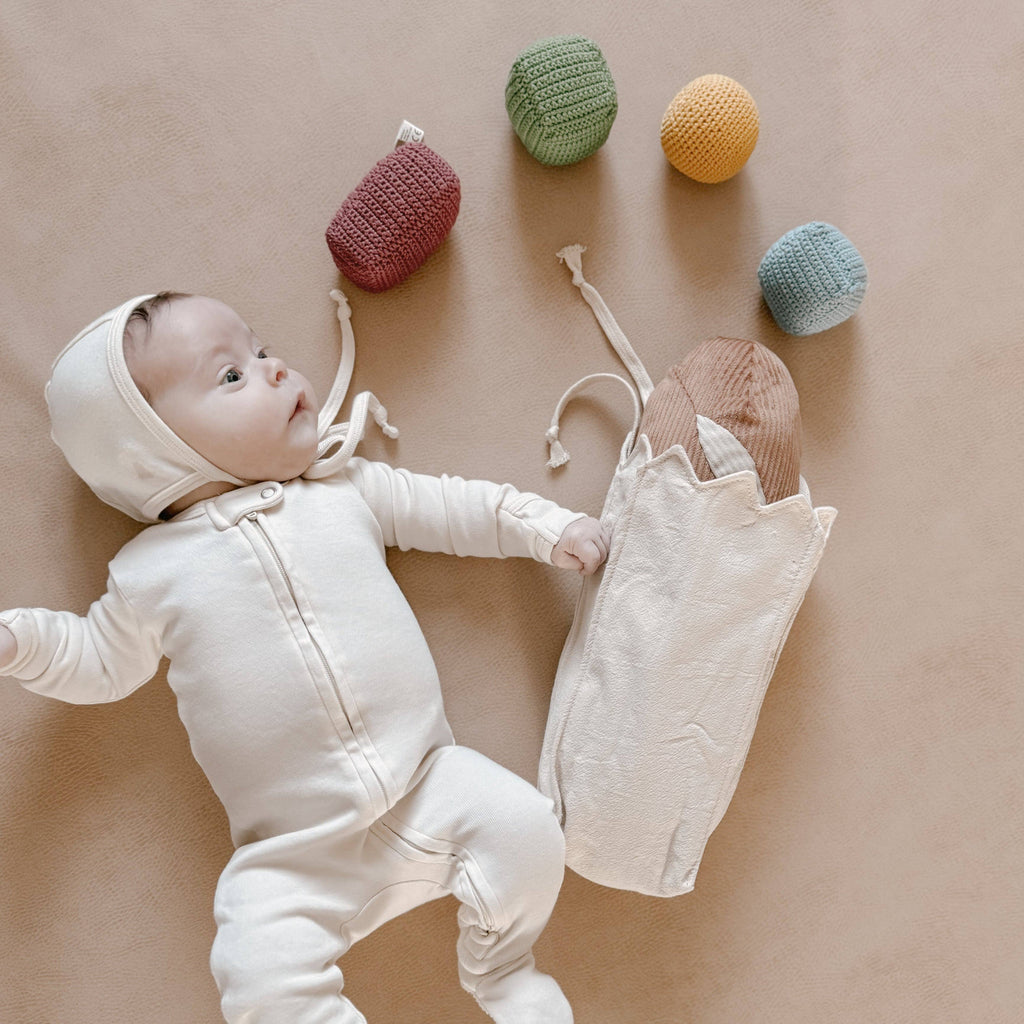 A baby in a cream-colored onesie and bonnet lies on a beige surface beside the Tummy Time Baguette—a drawstring bag filled with wooden toys, sensory beads, and four colorful crocheted balls for tummy time or pretend bakery fun.