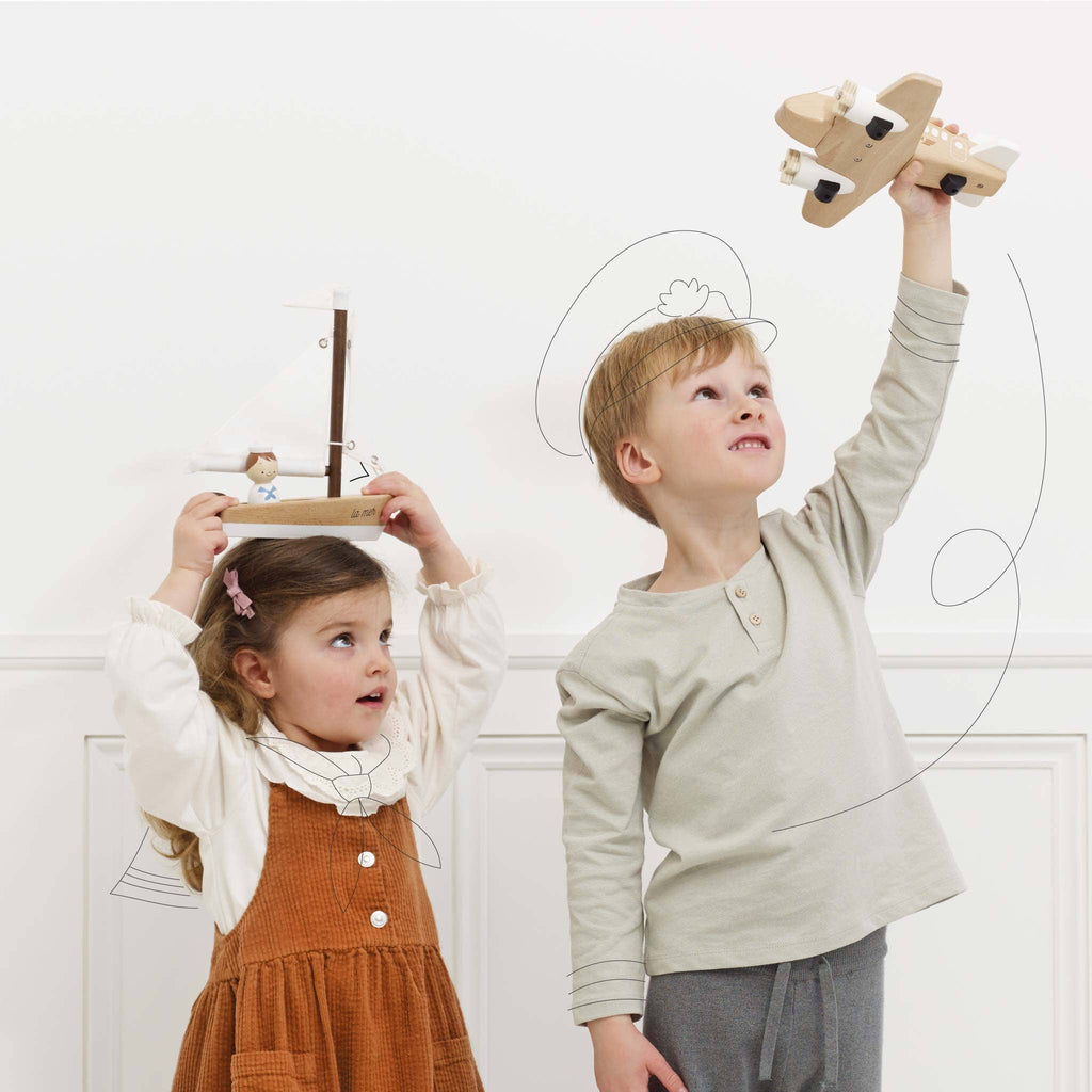 Two young children play indoors; the girl balances a wooden sailboat on her head while the boy lifts a Wooden Toy Plane above him. Both look up, smiling, surrounded by playful sketches of a boat and plane.