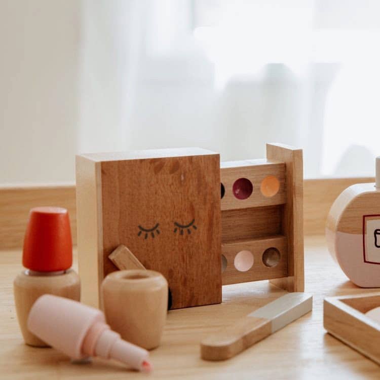The Wooden Beauty Kit sits on the table, complete with a nail polish, lipstick, smiley eyeshadow palette, and perfume bottle. Painted with non-toxic paint for safe play, it has a softly blurred background adding whimsy.