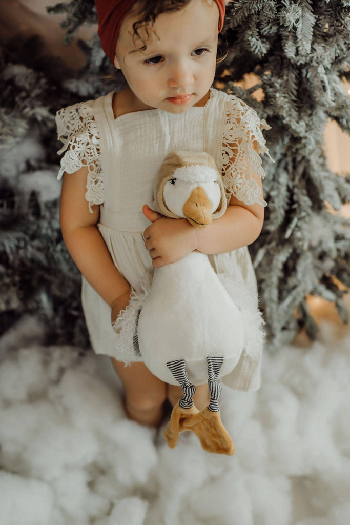 A young child in a white dress and red headband holds an Avery the Aviator Snow Goose Stuffed Animal while standing by a snow-covered pine tree. Faux snow is on the ground, and the child appears thoughtful.