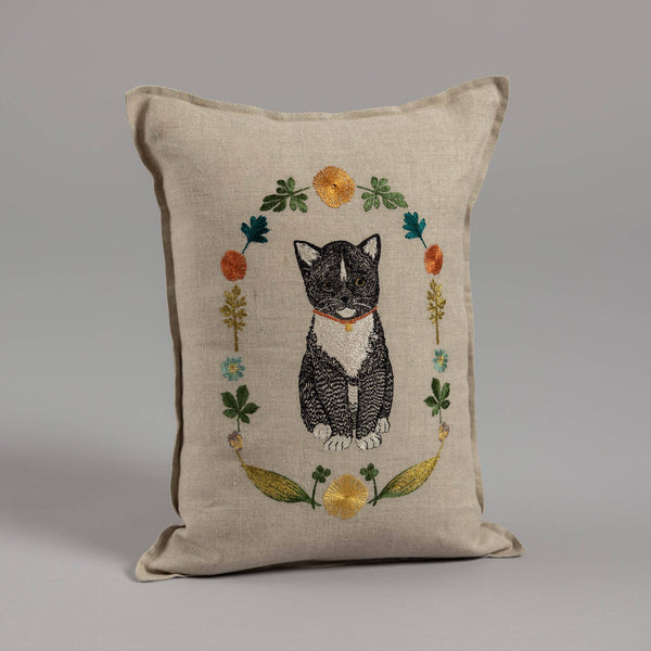 Introducing the Coral & Tusk Kitten Cameo Pillow: A beige cushion adorned with a black and white tuxedo cat with a red collar, surrounded by colorful spring flowers and leaves, on a plain gray background. Ships in one week.