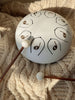 A Steel Tongue Drum, a unique musical instrument with marked notes, sits on a knitted cream blanket. Two wooden mallets rest nearby, bathed in gentle sunlight, creating a calm and inviting scene.