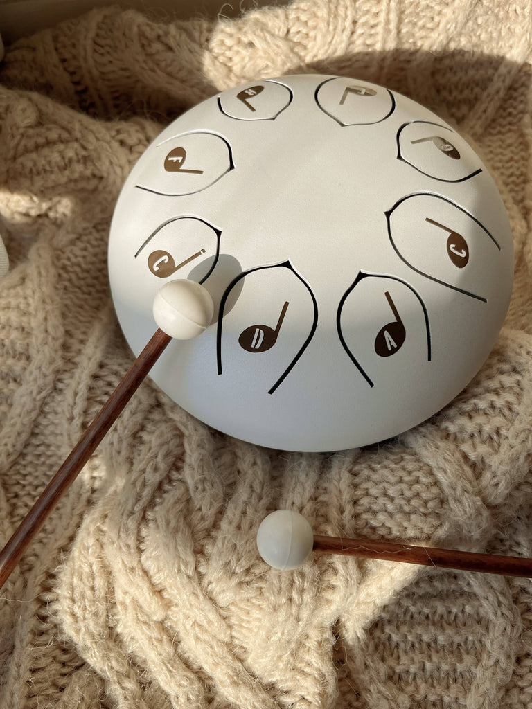 A Steel Tongue Drum, a unique musical instrument with marked notes, sits on a knitted cream blanket. Two wooden mallets rest nearby, bathed in gentle sunlight, creating a calm and inviting scene.