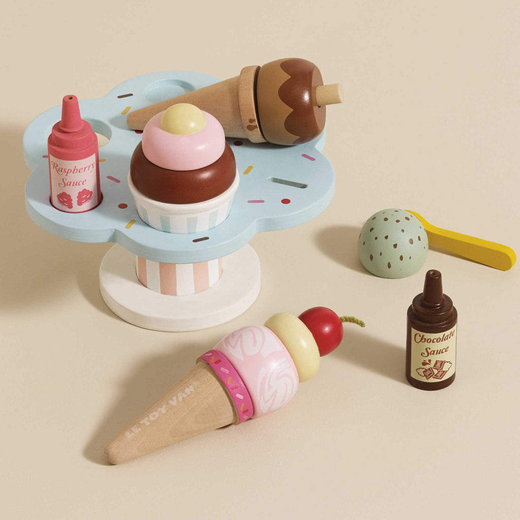 The Wooden Ice Cream Stand & Toppings set features colorful wooden toy desserts like ice cream cones and a cupcake on a blue sprinkle-patterned gelato stand, with raspberry and chocolate sauce bottles nearby, plus a mint chip scoop—ideal for role play fun.