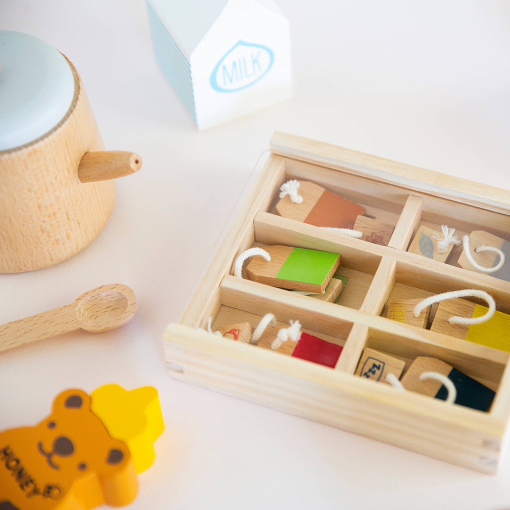 The Wooden Tea Set includes a wooden box filled with colorful food shapes, a charming teapot resembling an organic Matcha set, a yellow teddy bear piece, and a milk carton, all neatly arranged in their pretend play space on a white surface.