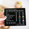 A hand holds a card from the Wooden Tea Set titled "Tea Orders Please!" with options like chai, mint, and organic Matcha Tea. Symbols for milk, honey, lemon, mint, and biscuits add a personal touch. It's like choosing your favorite tea in a cozy play space. Table number section sits atop the card.