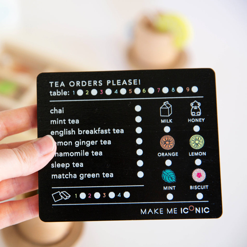 A hand holds a card from the Wooden Tea Set titled "Tea Orders Please!" with options like chai, mint, and organic Matcha Tea. Symbols for milk, honey, lemon, mint, and biscuits add a personal touch. It's like choosing your favorite tea in a cozy play space. Table number section sits atop the card.