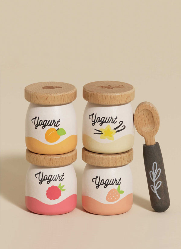 The Yogurt Play Food Pack contains four small containers with wooden lids stacked in two rows. Each container is labeled "Yogurt" and features fruit designs: orange, starfruit, raspberry, and strawberry. A small spoon with a decorative leaf pattern complements this plastic-free set.