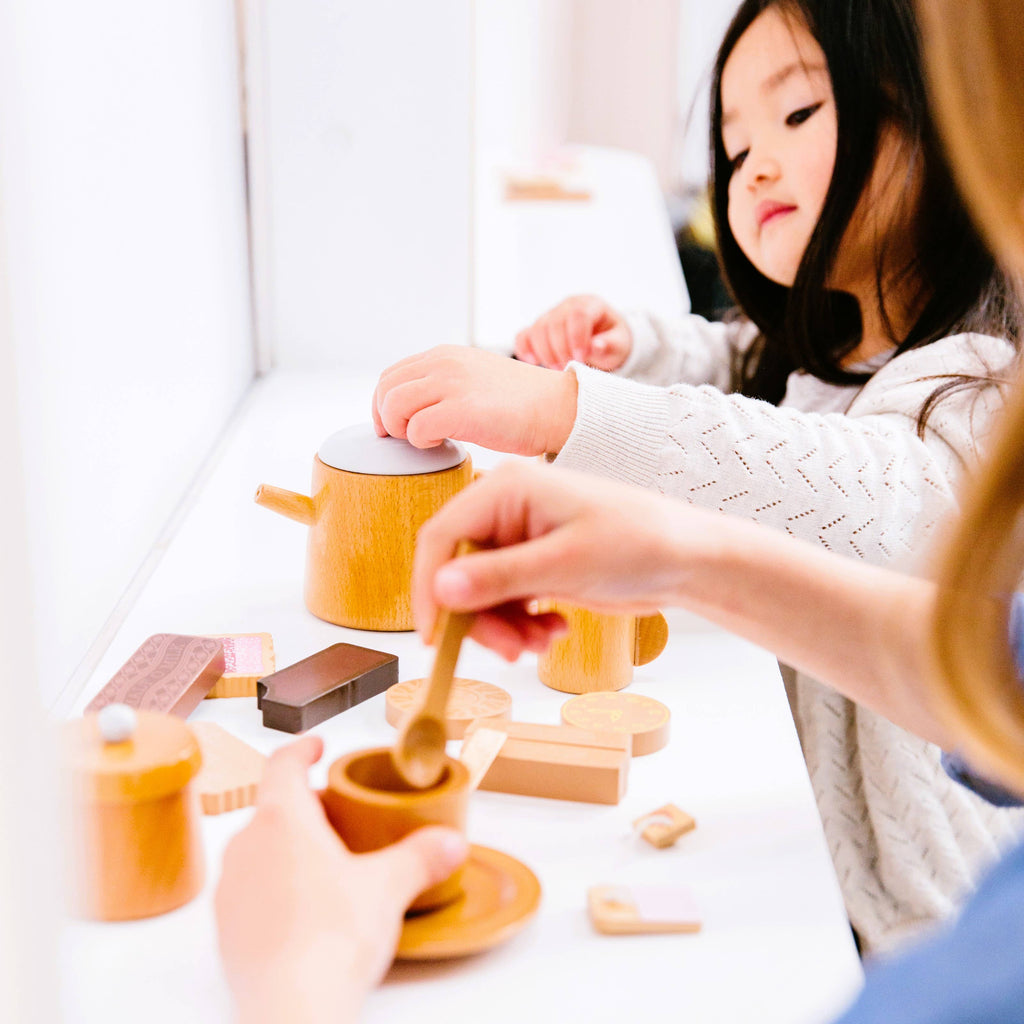 A child in a white sweater with long dark hair and an adult enjoy a warm, joyful moment mimicking chai latte making on a white surface using the Wooden Tea Set, featuring tiny pots, cups, and utensils.