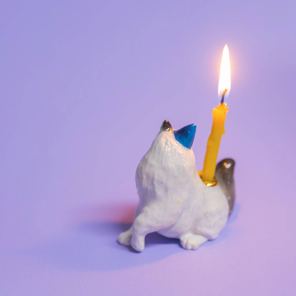 The Ragdoll Cat Cake Topper is a whimsical small, white ceramic wolf figurine howling with a lit yellow candle on its back. Against a light purple background, it adds an enchanting touch, perfect for cat-themed parties.