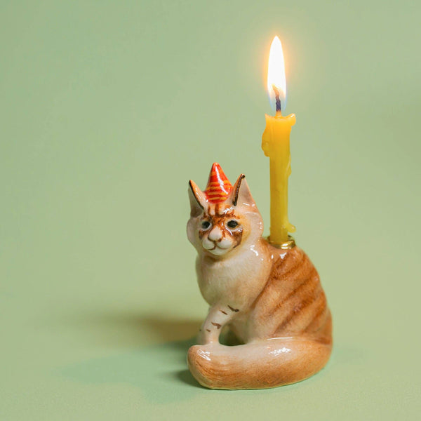 The Maine Coon Cat Cake Topper, featuring a striped design and party hat, rests on a light green backdrop. A lit yellow candle on its back emits a cozy glow, ideal for cat-themed celebrations.