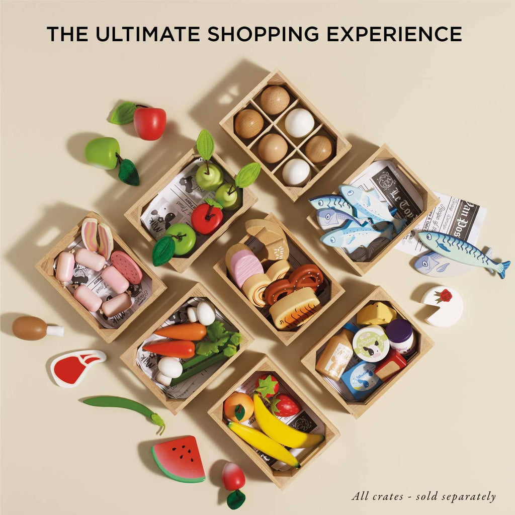 Displayed are toy grocery items, including role-play veggies, canned goods, fish, bread, and dairy. Wooden apples are neatly arranged in and around wooden crates on a pale background. Text above reads "The Ultimate Shopping Experience" featuring the Orchard Fruits Wooden Market Crate.
