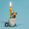 The Siamese Cat Cake Topper is a ceramic figurine, ideal for cat-themed parties, featuring a red polka-dotted party hat and carrying a lit, melted yellow candle on its back against a solid light blue background.