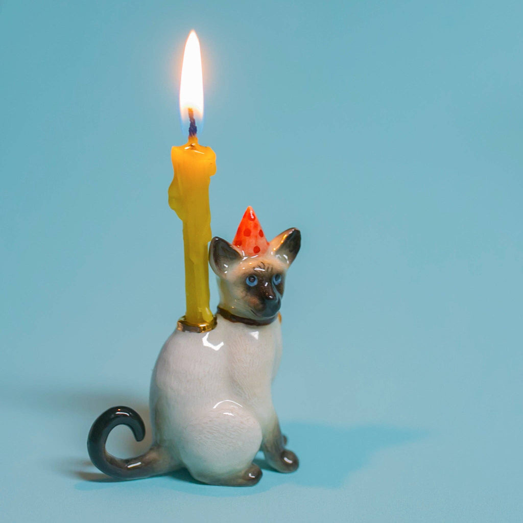 The Siamese Cat Cake Topper is a ceramic figurine, ideal for cat-themed parties, featuring a red polka-dotted party hat and carrying a lit, melted yellow candle on its back against a solid light blue background.