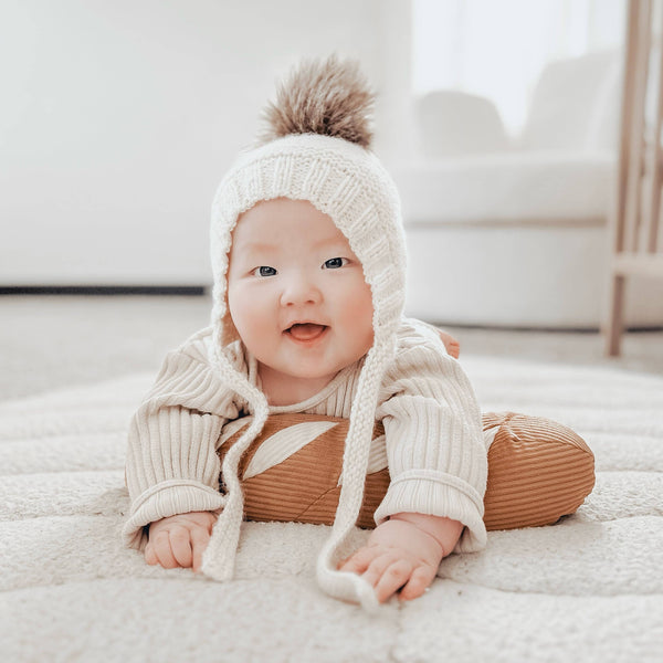 A smiling baby in a cream knit hat and ribbed outfit lies on a soft rug, gently resting on the Tummy Time Baguette pillow in a bright, cozy room.
