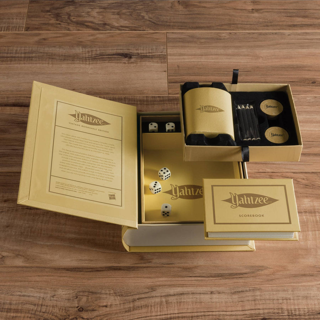 The Yahtzee Vintage Bookshelf Edition sits on a wooden surface, its elegant gold box open to display dice, scorebook, pencils, chips, and a dice cup—perfectly arranged for fans of classic dice games.