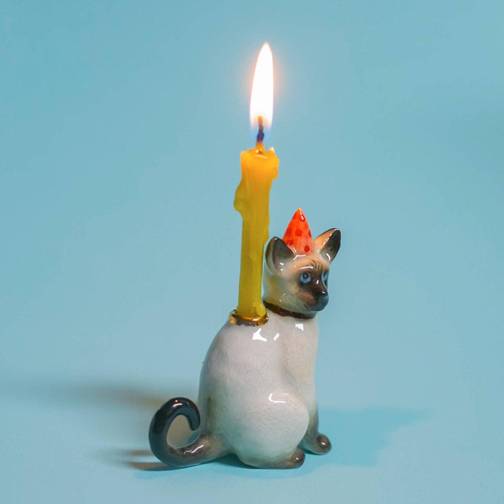 The Siamese Cat Cake Topper is a ceramic figurine of a Siamese cat in a red party hat, with a lit yellow candle on its back. Ideal for whimsical cat-themed parties, it features a light blue background and casts a small shadow from the candle.