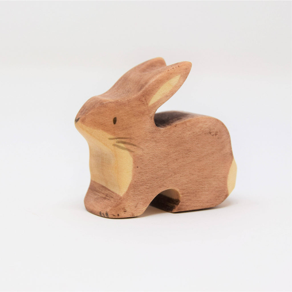 The Handmade Wooden Brown Rabbit is a light brown, hand-crafted wooden figurine with simple carved details for eyes, ears, and whiskers. Made from sustainable hardwood and enhanced with plant-based stains, the rabbit faces left against a plain white background.