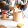 A child delightfully plays with the Wooden Tea Set, pretending it's filled with almond chai. On a white table are the teapot and teacup with saucer. Wearing a gray sweater with colorful patches, the child is immersed in an imaginary tea party world.