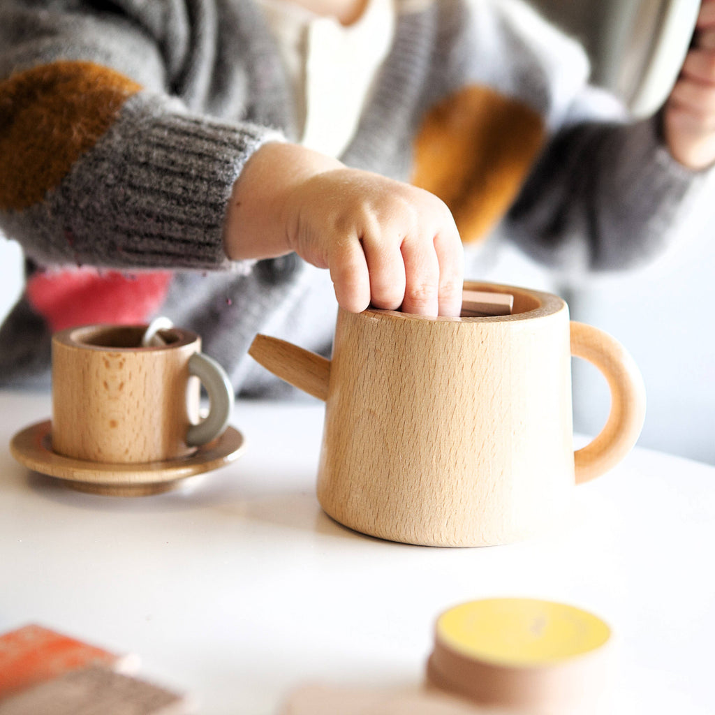 A child delightfully plays with the Wooden Tea Set, pretending it's filled with almond chai. On a white table are the teapot and teacup with saucer. Wearing a gray sweater with colorful patches, the child is immersed in an imaginary tea party world.