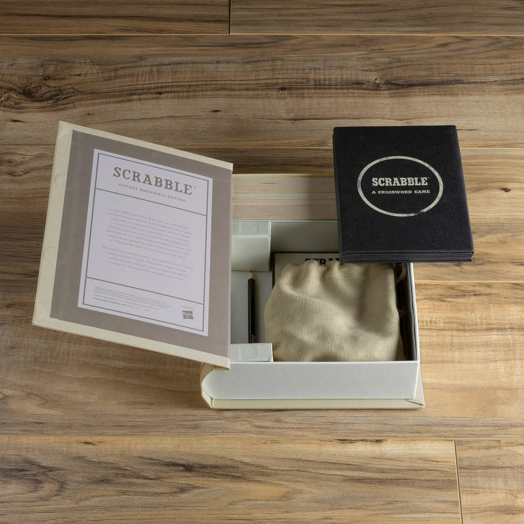 The Scrabble Vintage Bookshelf Edition box sits open on a wooden surface, revealing the rule book, pouch, and premium components inside. The lid, featuring "Scrabble: A Crossword Game," is propped open to the right.