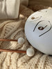 A Steel Tongue Drum, finished in white with note symbols, rests on a cream knitted blanket. Two wooden mallets with white rubber tips lie nearby, bathed in warm sunlight for a cozy look.