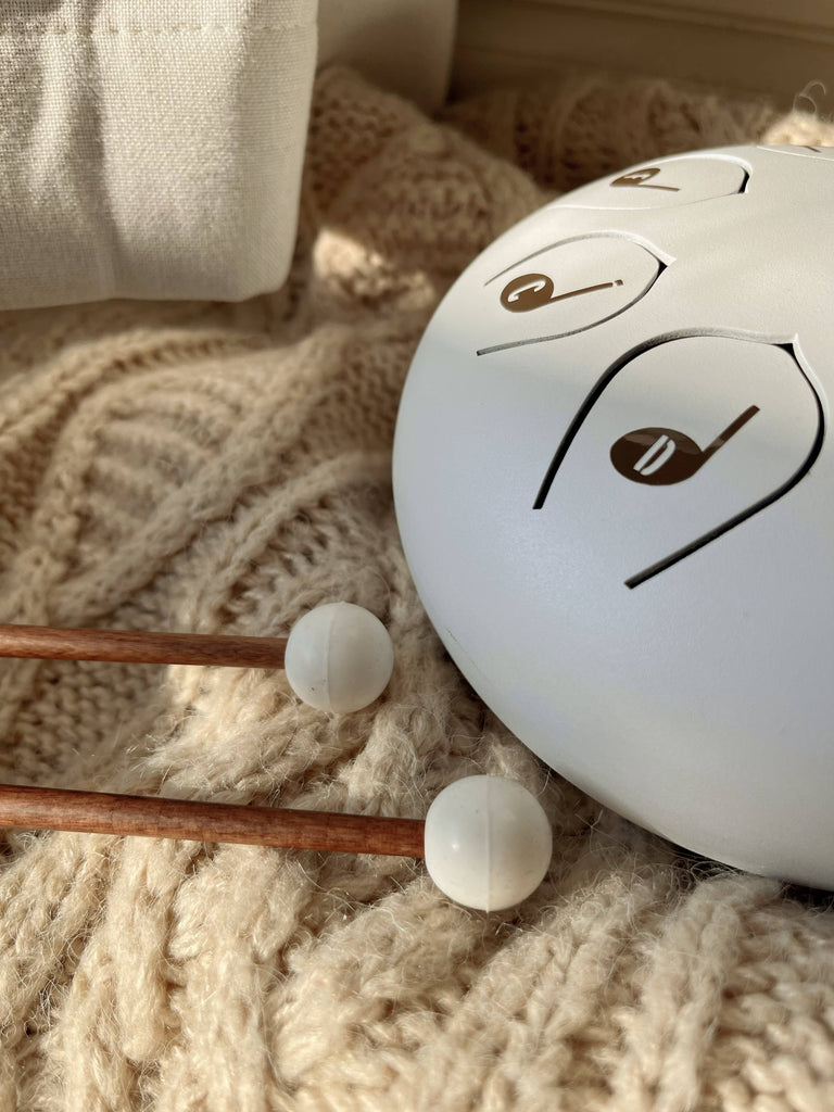 A Steel Tongue Drum, finished in white with note symbols, rests on a cream knitted blanket. Two wooden mallets with white rubber tips lie nearby, bathed in warm sunlight for a cozy look.