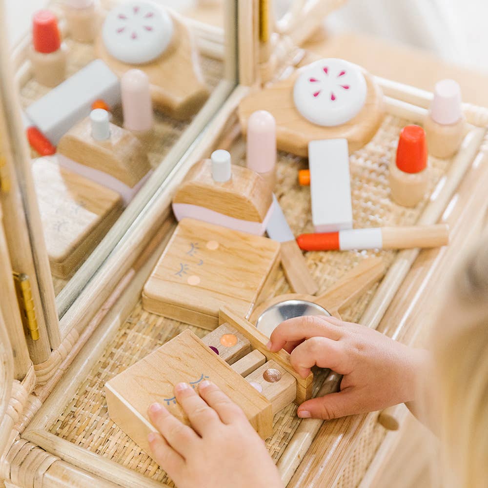 A child explores a timeless toy, the Wooden Beauty Kit, featuring pastel-colored brushes and compacts. Crafted with non-toxic paint, it sits elegantly on a wicker shelf by a mirror, sparking playful imagination.