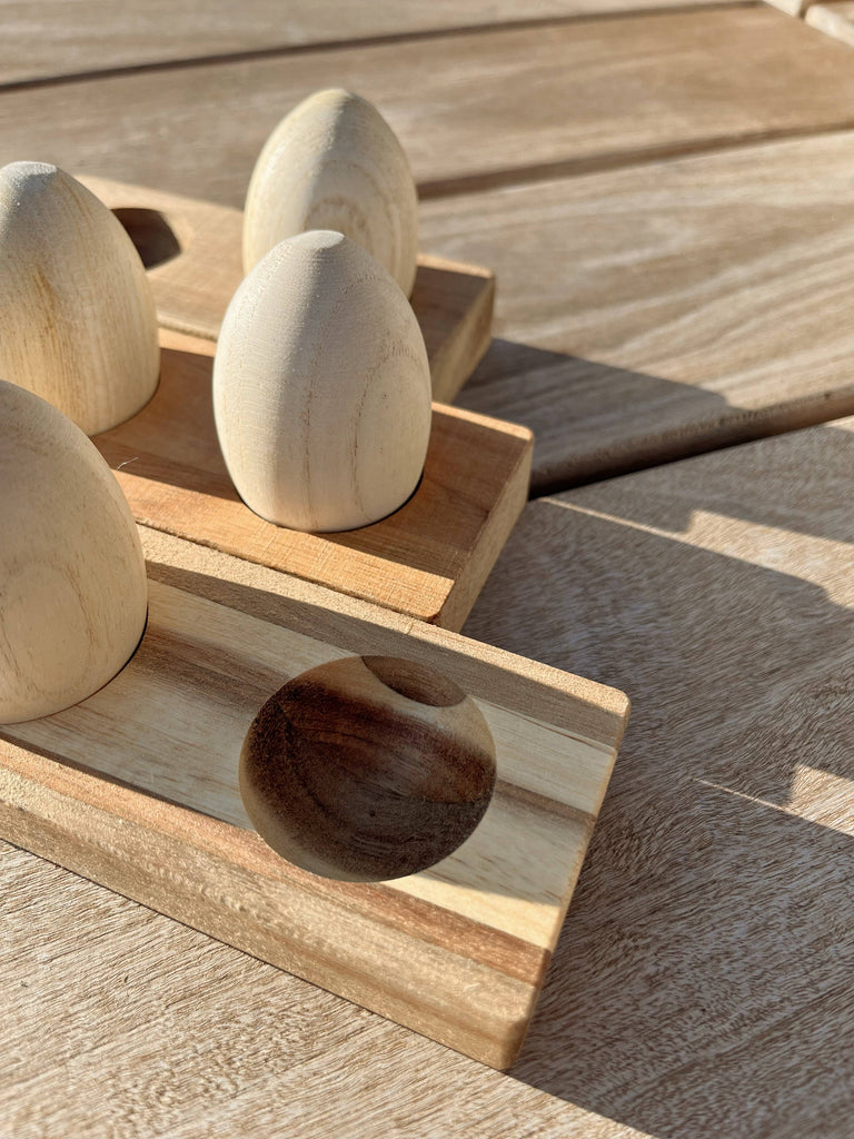 Four Wooden Egg Shakers with Stand—smooth, egg-shaped, and placed in indented slots on a natural wood tray—rest on a sunlit wooden surface, one slot empty and soft shadows cast around them.