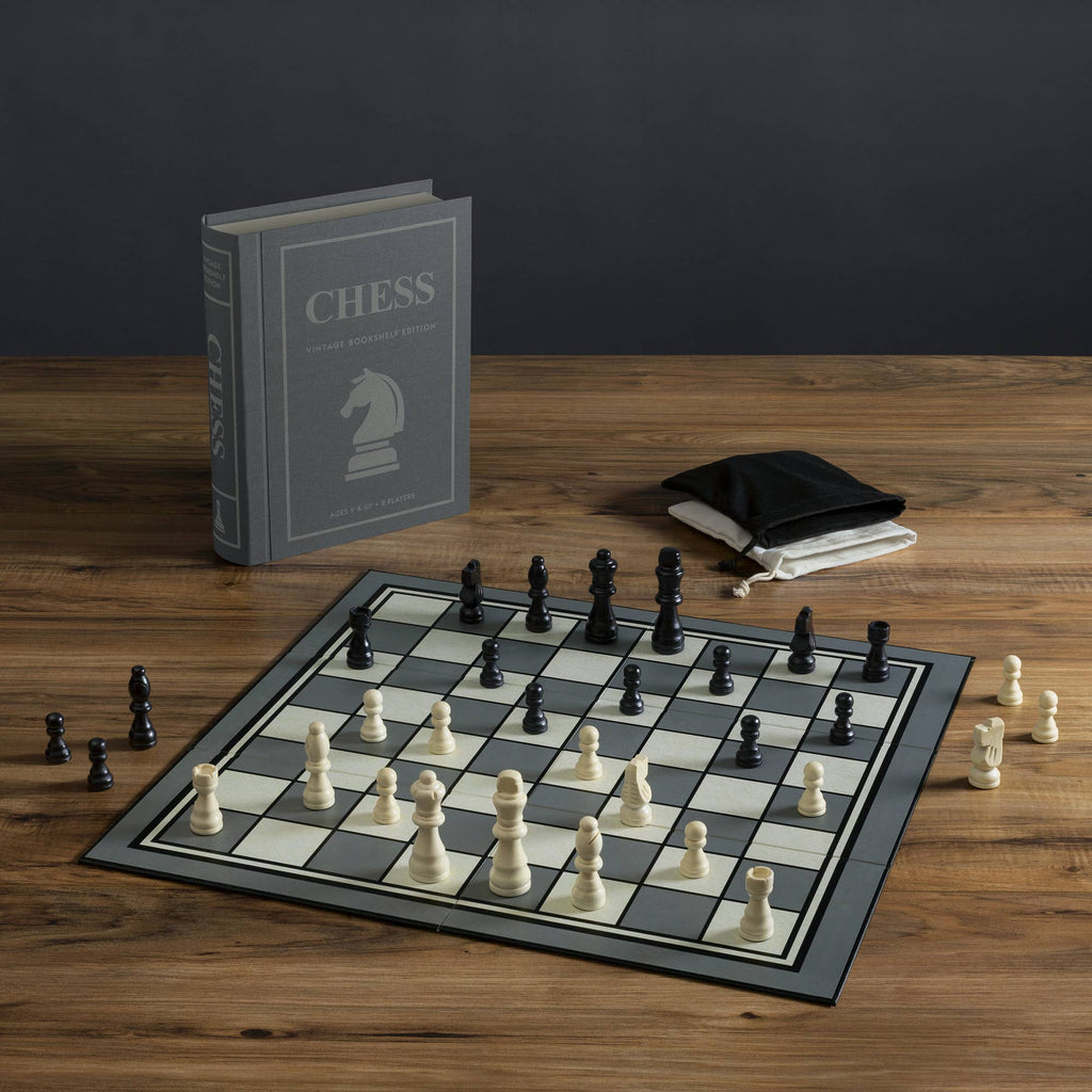 Chess Vintage Bookshelf Edition: A classic strategy game featuring white and black pieces on a board, a standing chess instruction book, and two drawstring bags—all elegantly arranged for collectors.