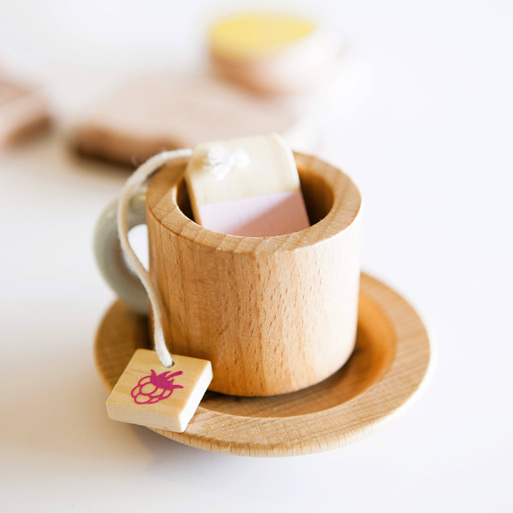 The Wooden Tea Set includes a light brown wooden toy tea cup with a saucer and a small wooden bag featuring a string and pink tag resembling a tea bag, perfect for creating a mini chai latte vibe. The background has blurred objects for added ambiance.