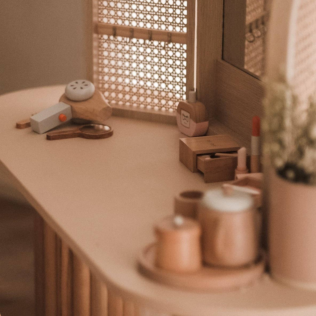 A cozy vanity setup with a wooden table includes beauty products like the Wooden Beauty Kit, perfumes, lipstick, and a small wooden box. A mirror with a lattice pattern and camera-shaped decor add charm, while soft lighting enhances the scene's warmth.