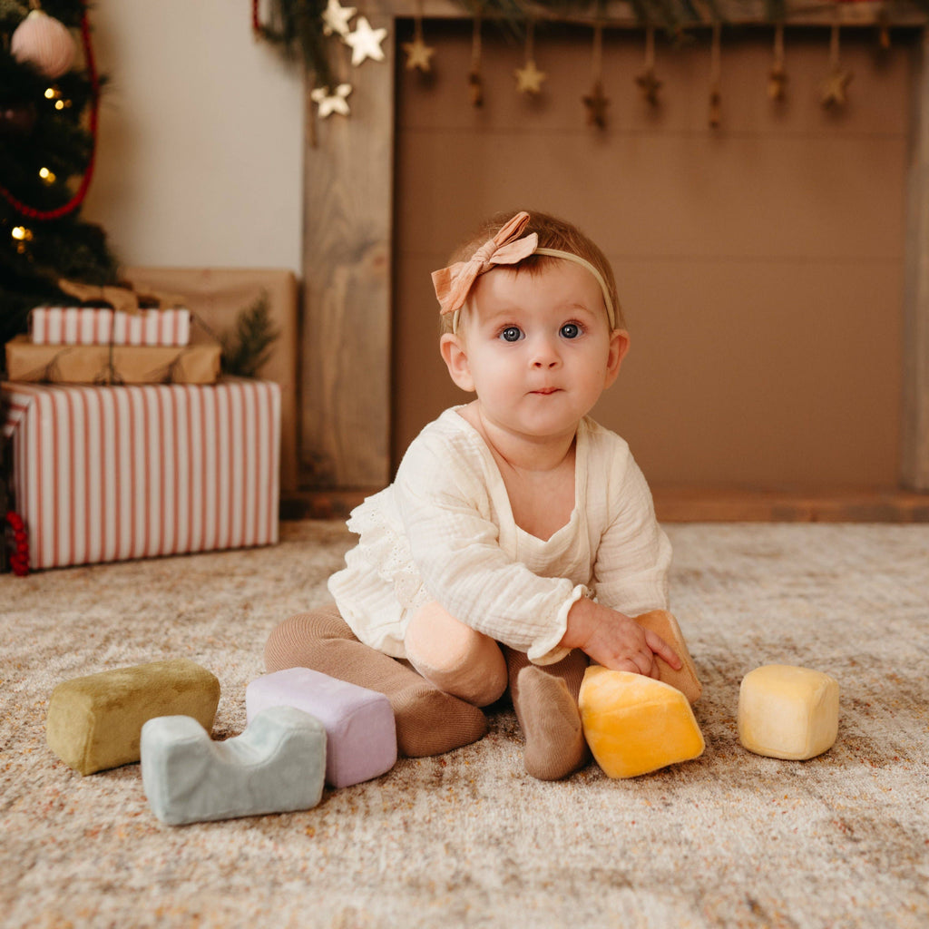 A baby in a cream outfit with a brown bow sits on a carpet, playing with Soft Toy Blocks. Wrapped gifts and a decorated Christmas tree are in the background, creating a cozy holiday atmosphere.
