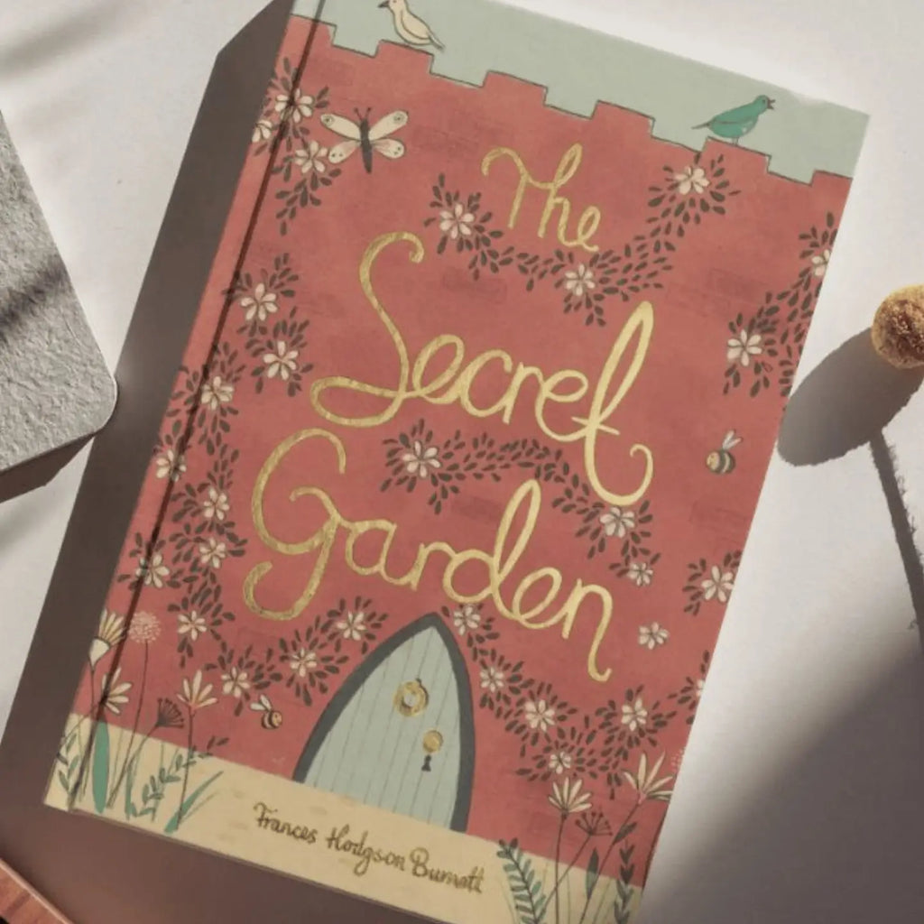 The Secret Garden | Burnett | Collector’s Edition | Hardcover rests on a light surface, its pink cover adorned with floral designs, bees, birds, and a small illustrated door at the bottom.