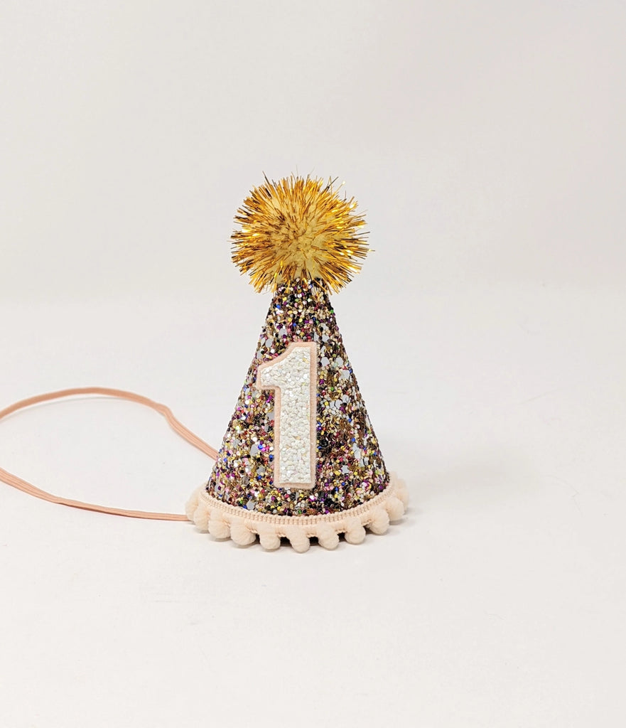 The Confetti Glitter Party Hat is a small, conical party hat with a gold glittery surface adorned with multicolored sparkles, perfect for birthday celebrations and photo shoots. The hat has a gold pom-pom on top, a white number 1 on the front, white pom-pom trim around the base, and a light pink elastic chin strap.