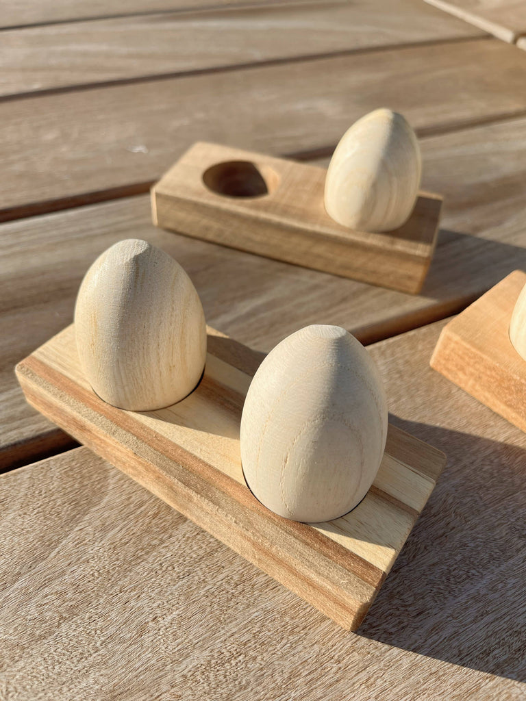 Two Wooden Egg Shakers with Stand sit on a rectangular wooden base, with another set behind them, all arranged on a sunlit light wood surface.